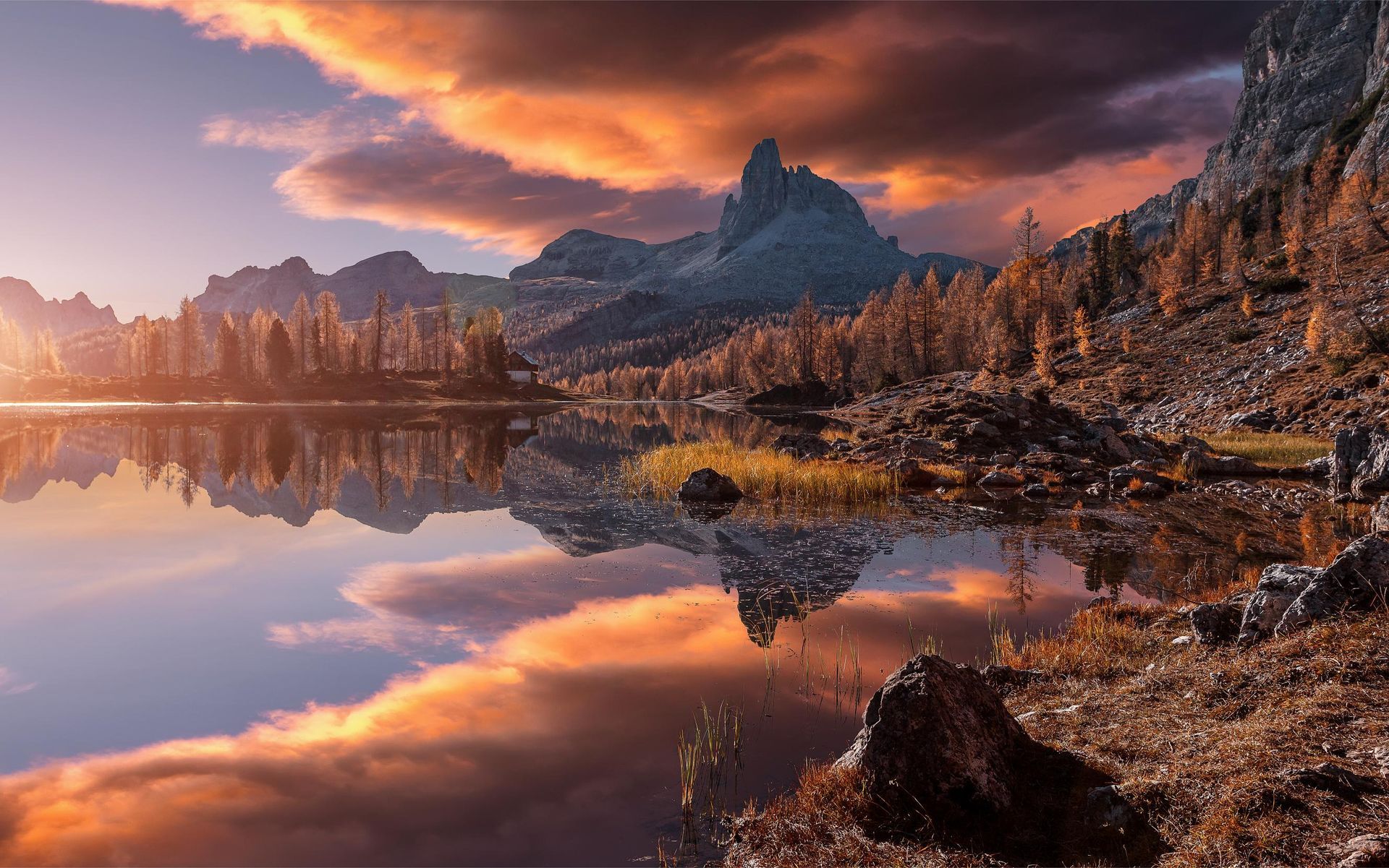 A golden sunset glows over a calm mountain lake, reflecting the jagged peaks and orange clouds in the still water.