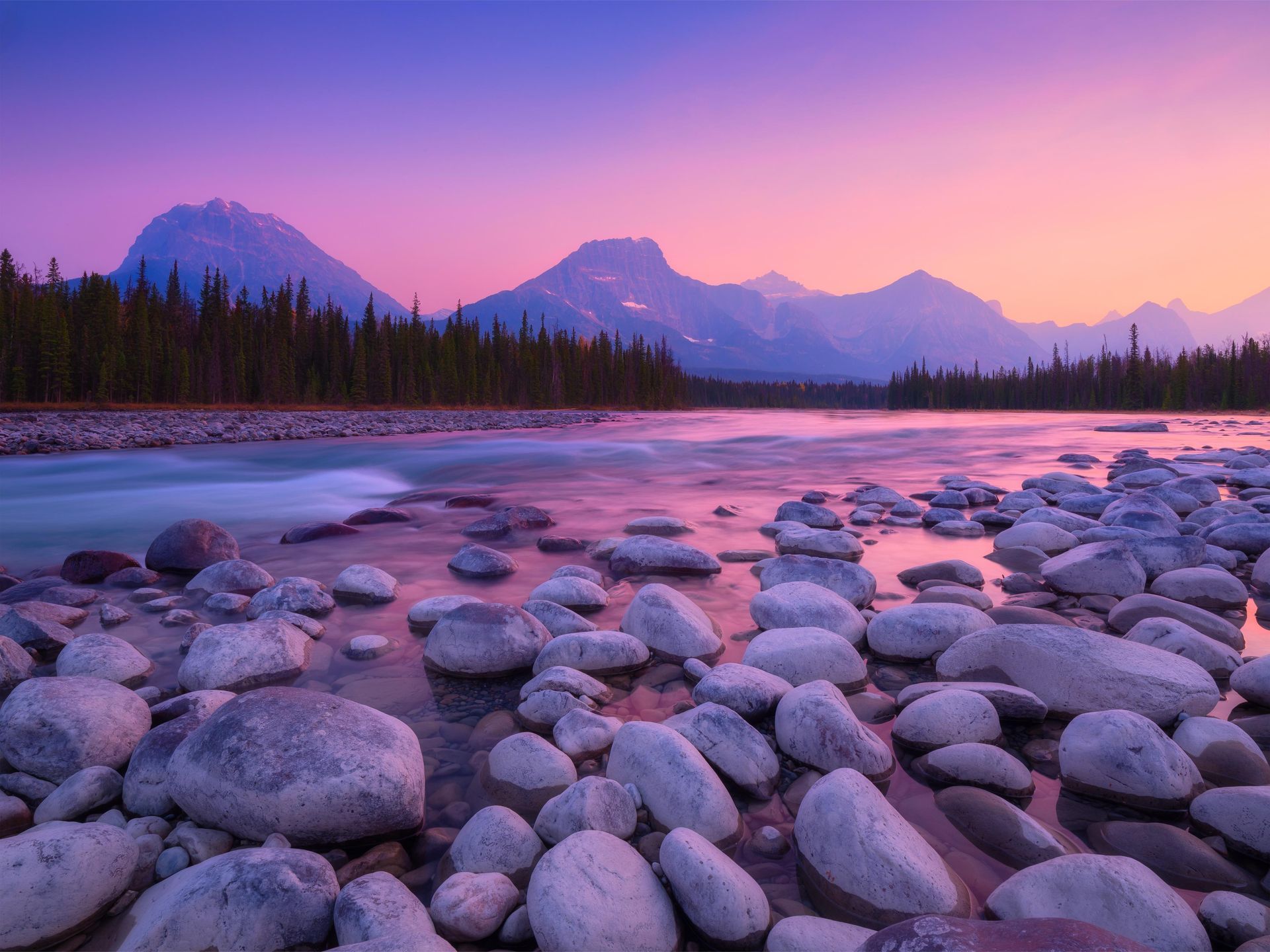 A rocky riverbank at sunset with smooth gray stones in the foreground and a mountain range against a purple sky.
