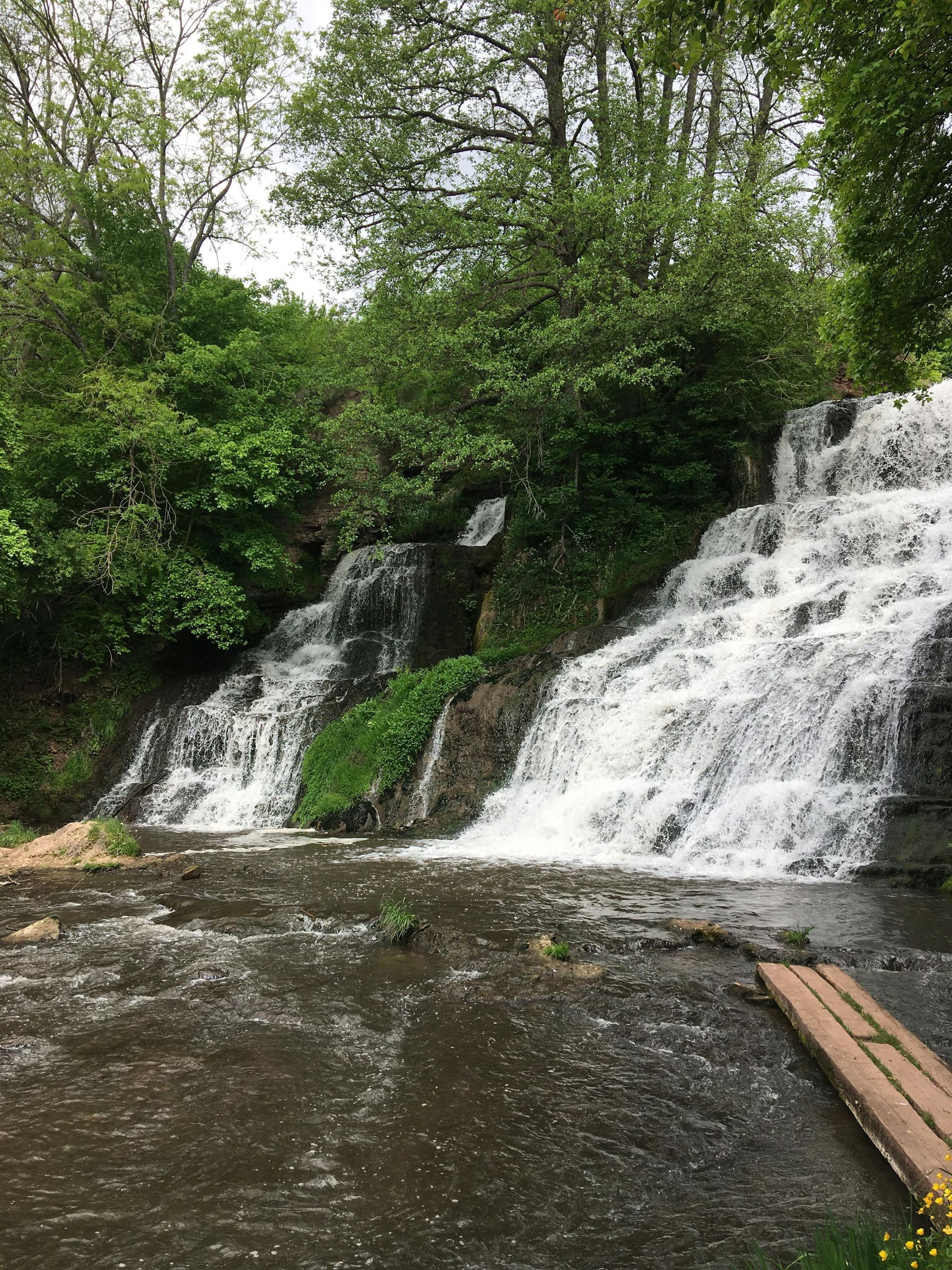 Two waterfalls cascade down rocky, tree-lined slopes into a dark pool, with a wooden boardwalk in the foreground.