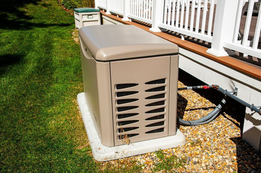 Tan residential generator next to a house on a concrete pad with vent. Green lawn.