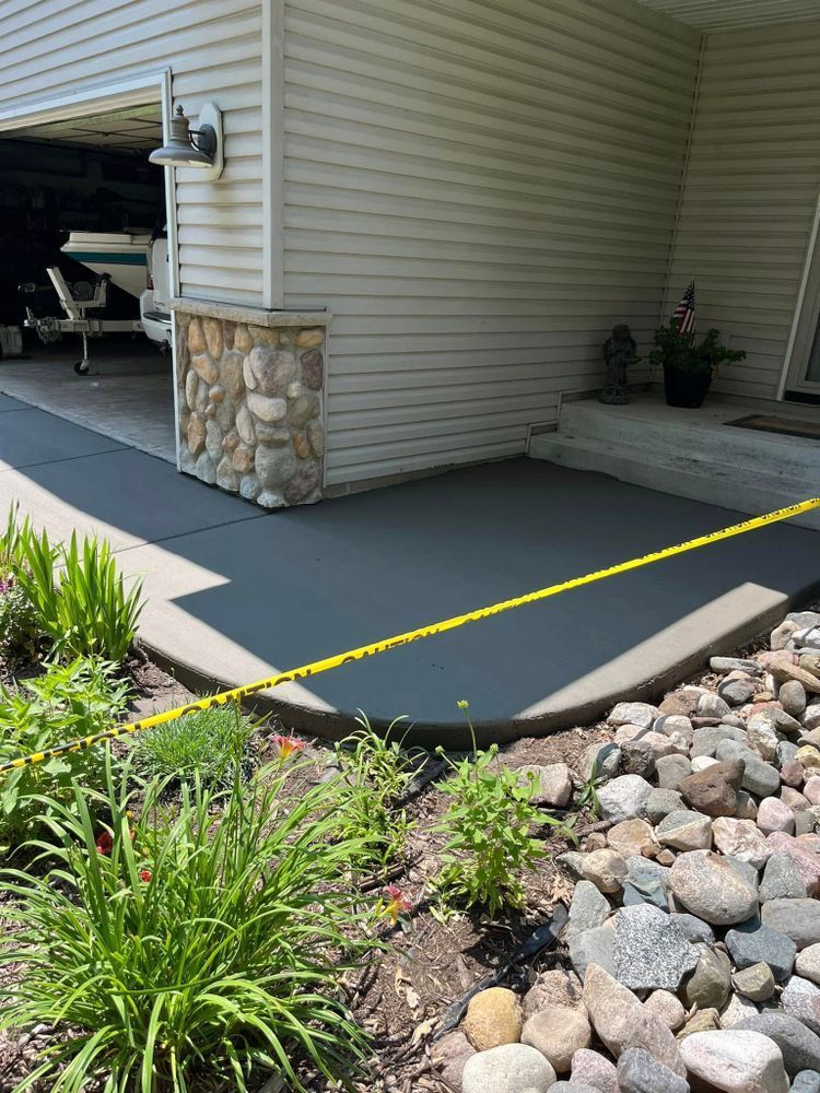 Freshly poured gray concrete walkway next to a house with a stone-covered pillar; yellow caution tape is present.