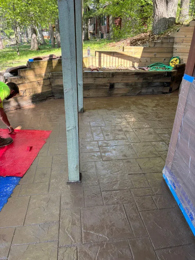 Man applying sealant to a patterned concrete patio. Green and blue mats, wooden support beams. Outdoor setting.