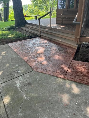 Concrete patio with textured, reddish-brown section next to a wooden deck with steps and railing.