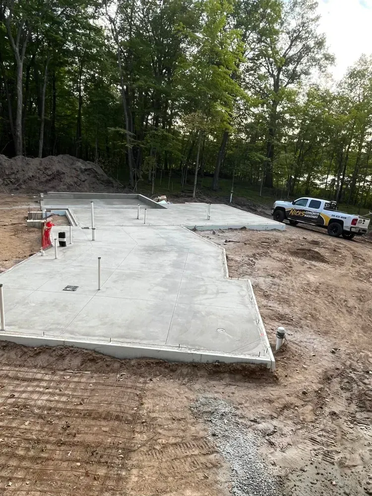 Concrete foundation of a building under construction, with plumbing and a truck parked nearby. Forest background.