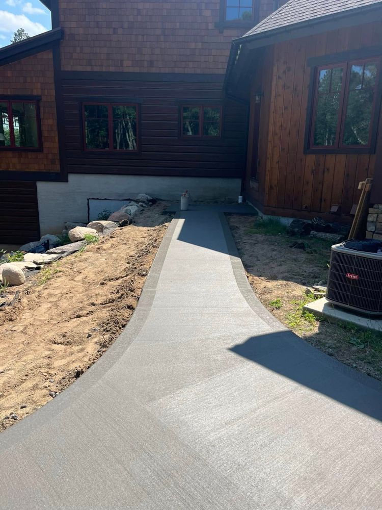 Concrete walkway leading to a brown wood-sided house. Pathway borders dirt and lawn.
