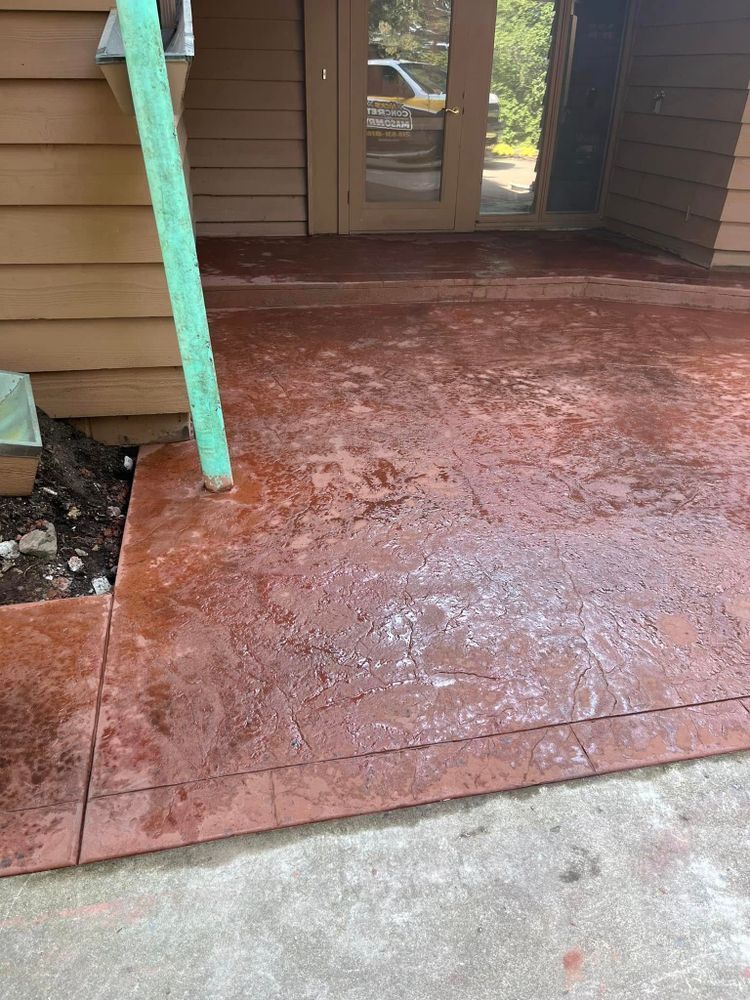 Stamped concrete patio with a red-brown hue, wet with water, next to a building with brown siding and a doorway.