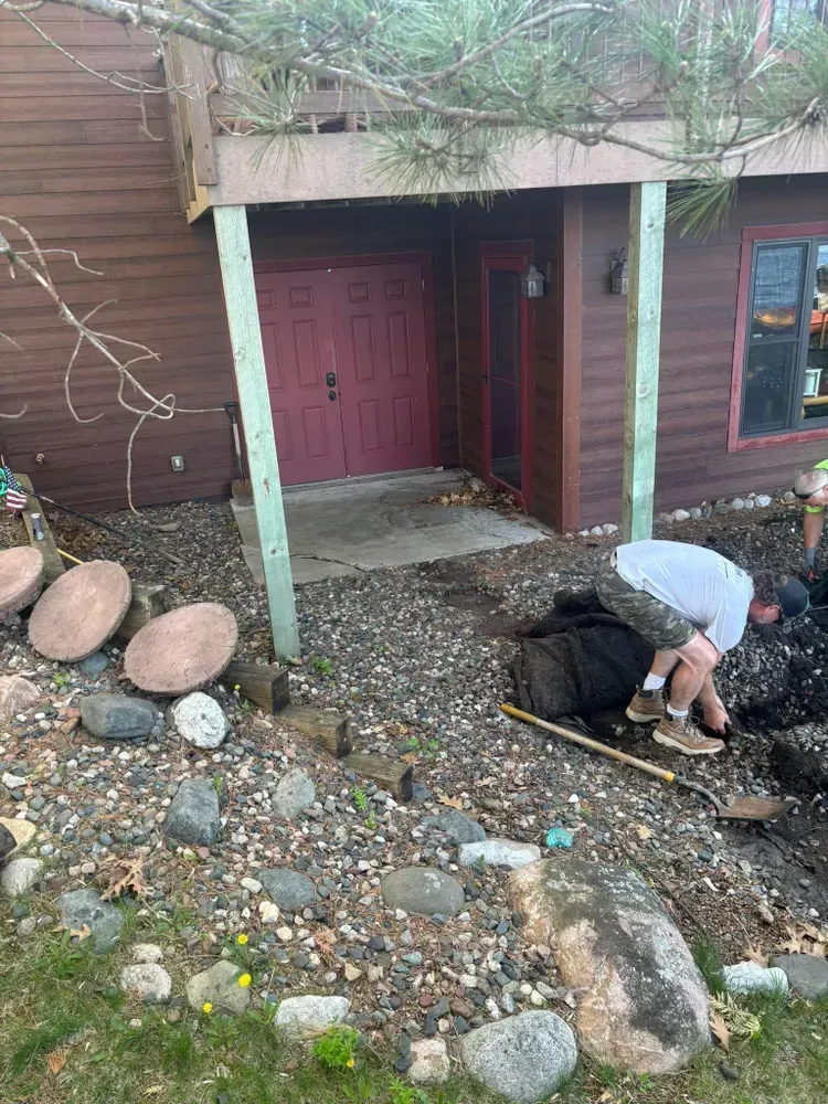 Man digging in a garden bed near a house entrance with rocks and stepping stones.