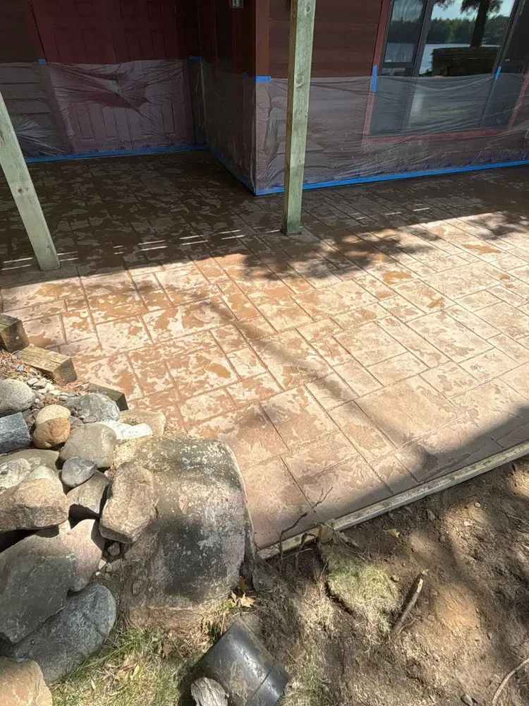 Concrete patio with brick pattern, brown color, near rocks and building with plastic wrap.