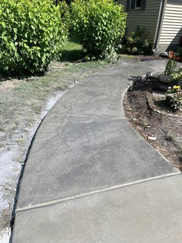 Curving concrete walkway in a yard with green bushes and grass, leading to a house.