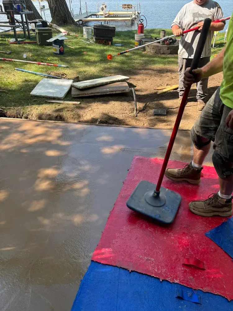 Person smoothing wet concrete with a tool, on a red and blue surface outdoors near water.