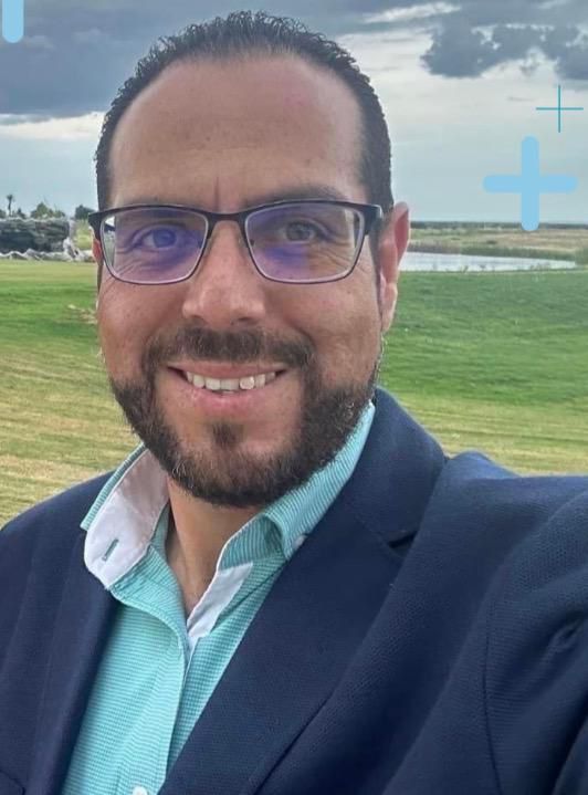 Un hombre con gafas sonríe, vistiendo una chaqueta azul y una camisa verde azulado, al aire libre frente a un paisaje verde.