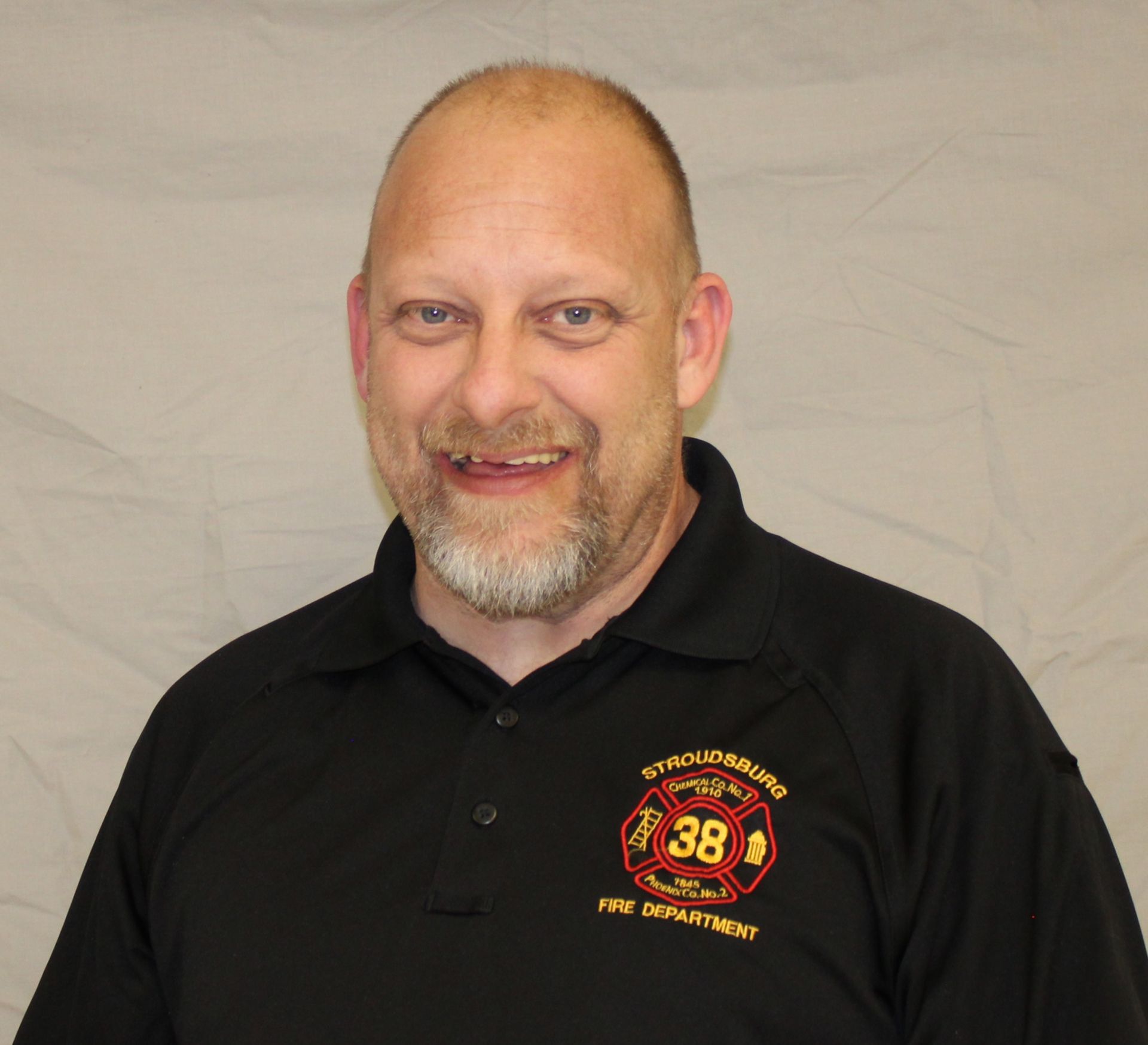 Man with short hair and beard in a black polo shirt with a fire department logo. He is smiling slightly.