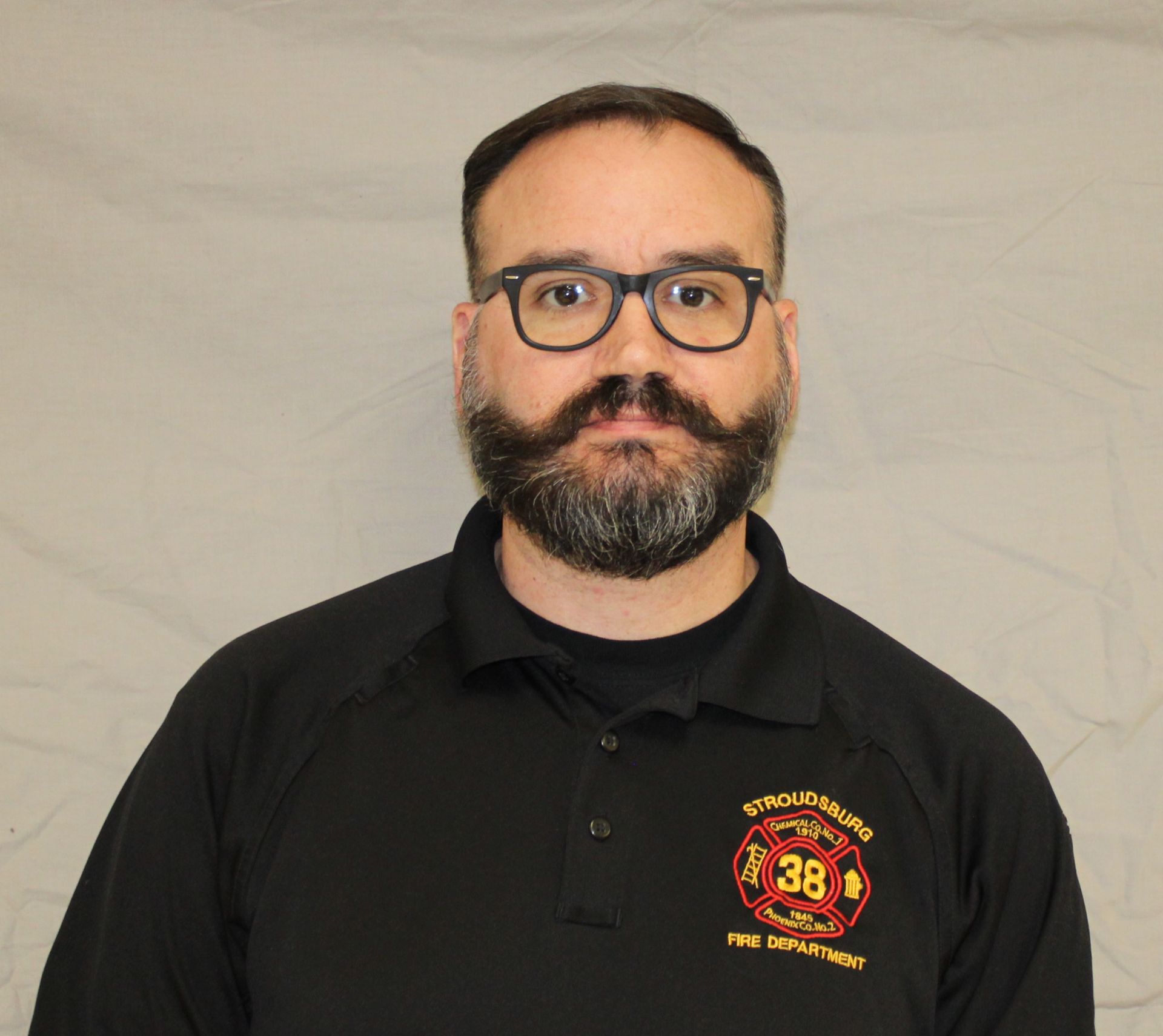 A man with a beard and glasses in a black shirt with a fire department logo. He looks straight ahead.