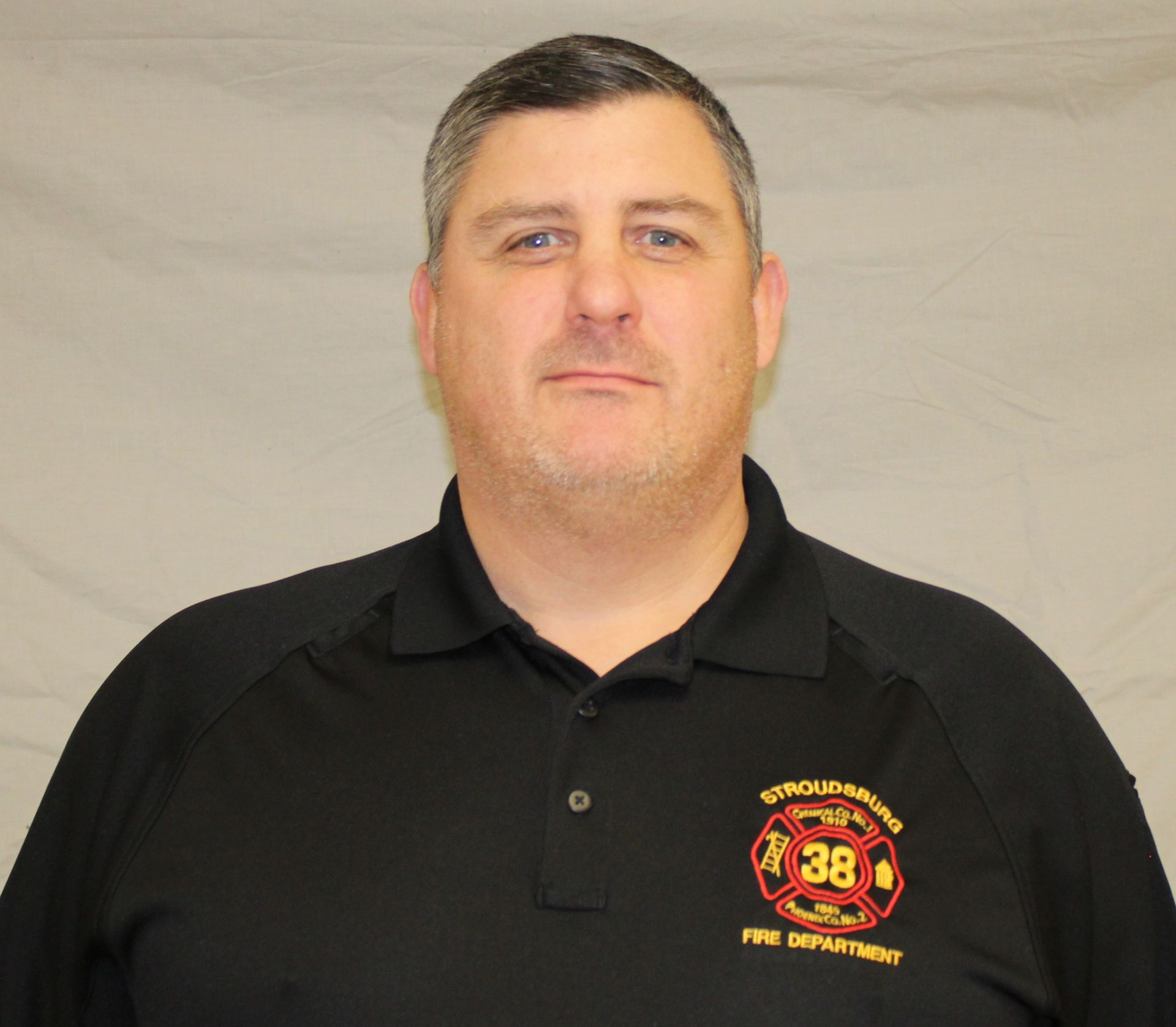 Man in a black polo shirt with a fire department emblem, against a plain backdrop.