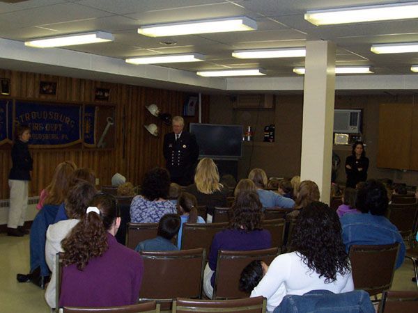 A firefighter speaks to a group of seated adults in a room. Two other people stand nearby. The room has wooden paneling and fluorescent lights.