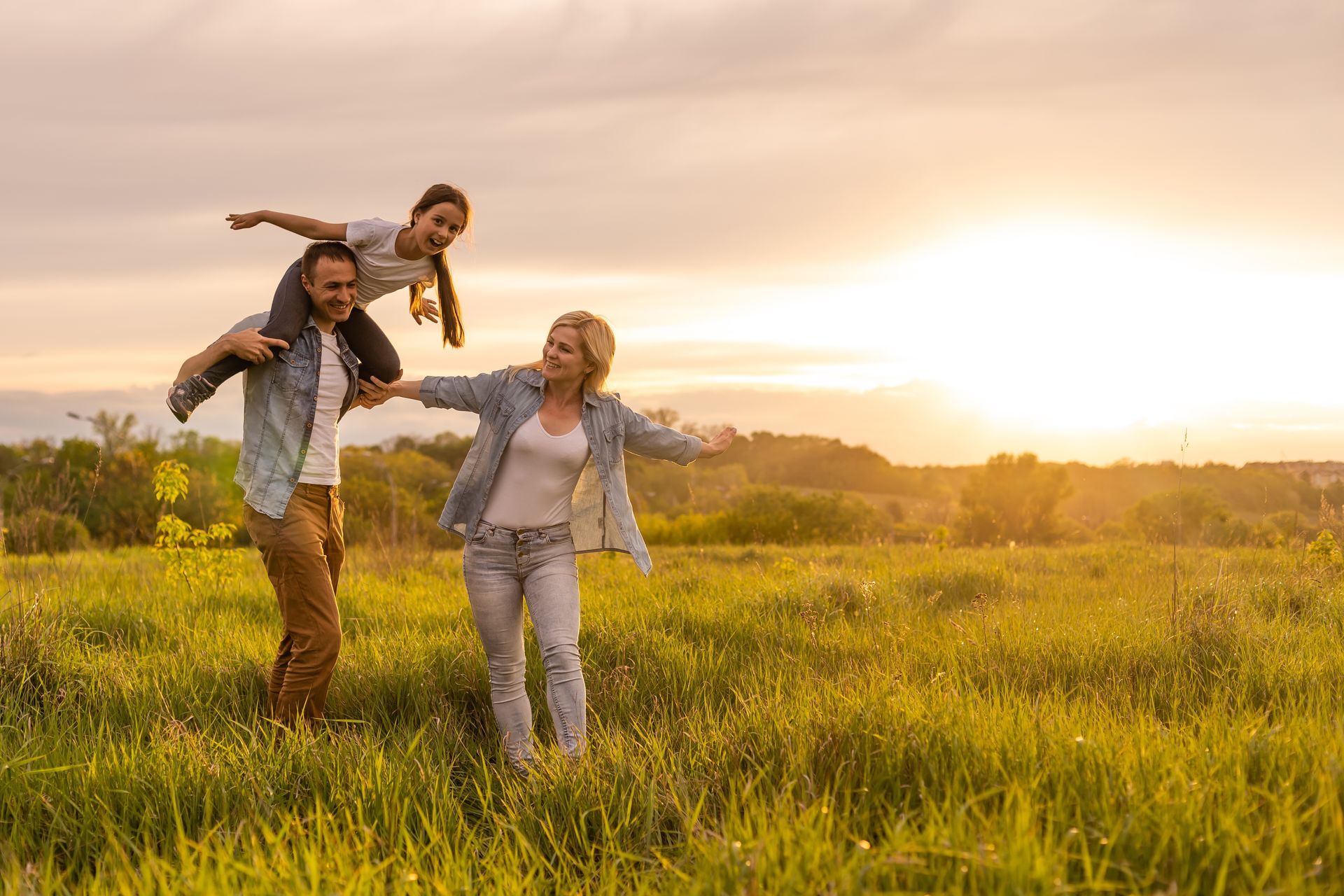 Family of three smiling outdoors: child on father's shoulders, mother smiling at them.