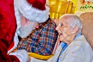 An elderly woman is sitting in a chair talking to santa claus.