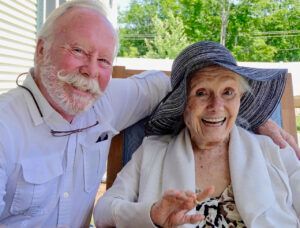 A man and an elderly woman are sitting next to each other on a porch . the woman is wearing a hat.