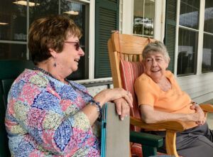 Two older women are sitting in rocking chairs on a porch.