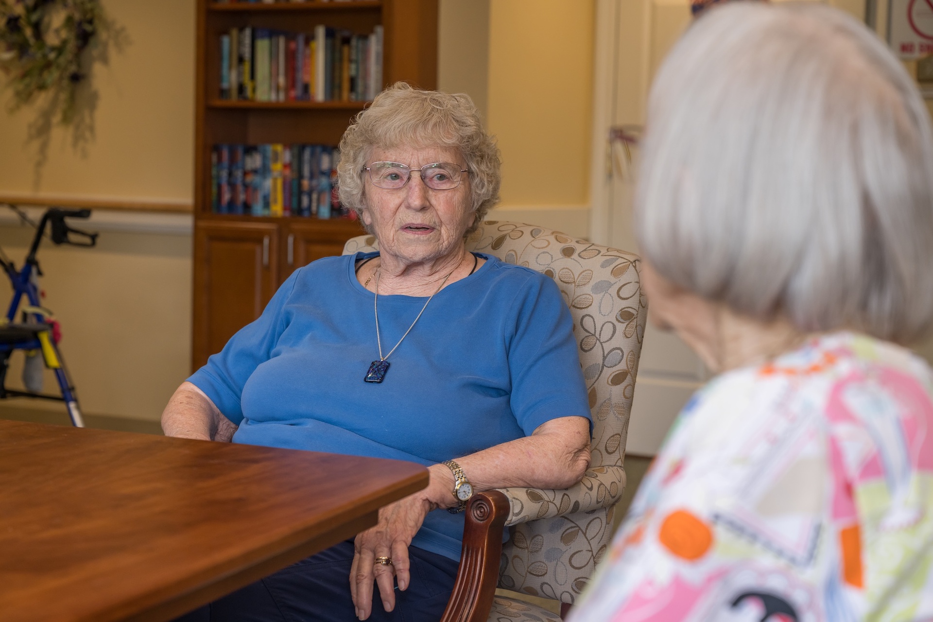 An older woman in a blue shirt sits at a table, looking towards another person.
