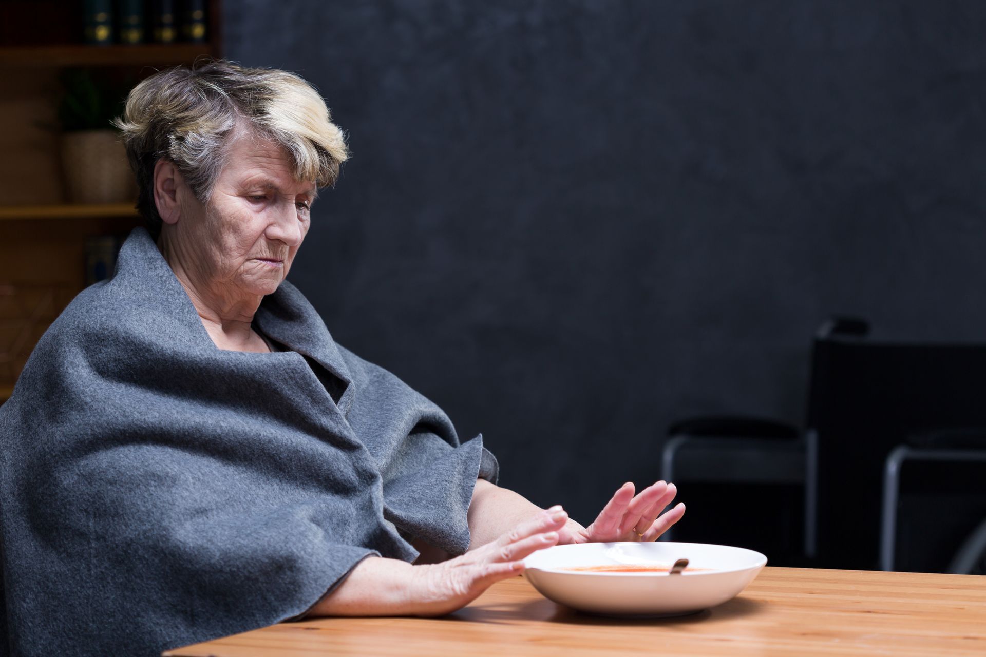An elderly woman wrapped in a blanket is sitting at a table with a bowl of soup.