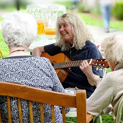 A woman is playing a guitar to two older women while sitting on a bench.