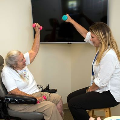 An elderly woman in a wheelchair is being helped by a nurse with dumbbells