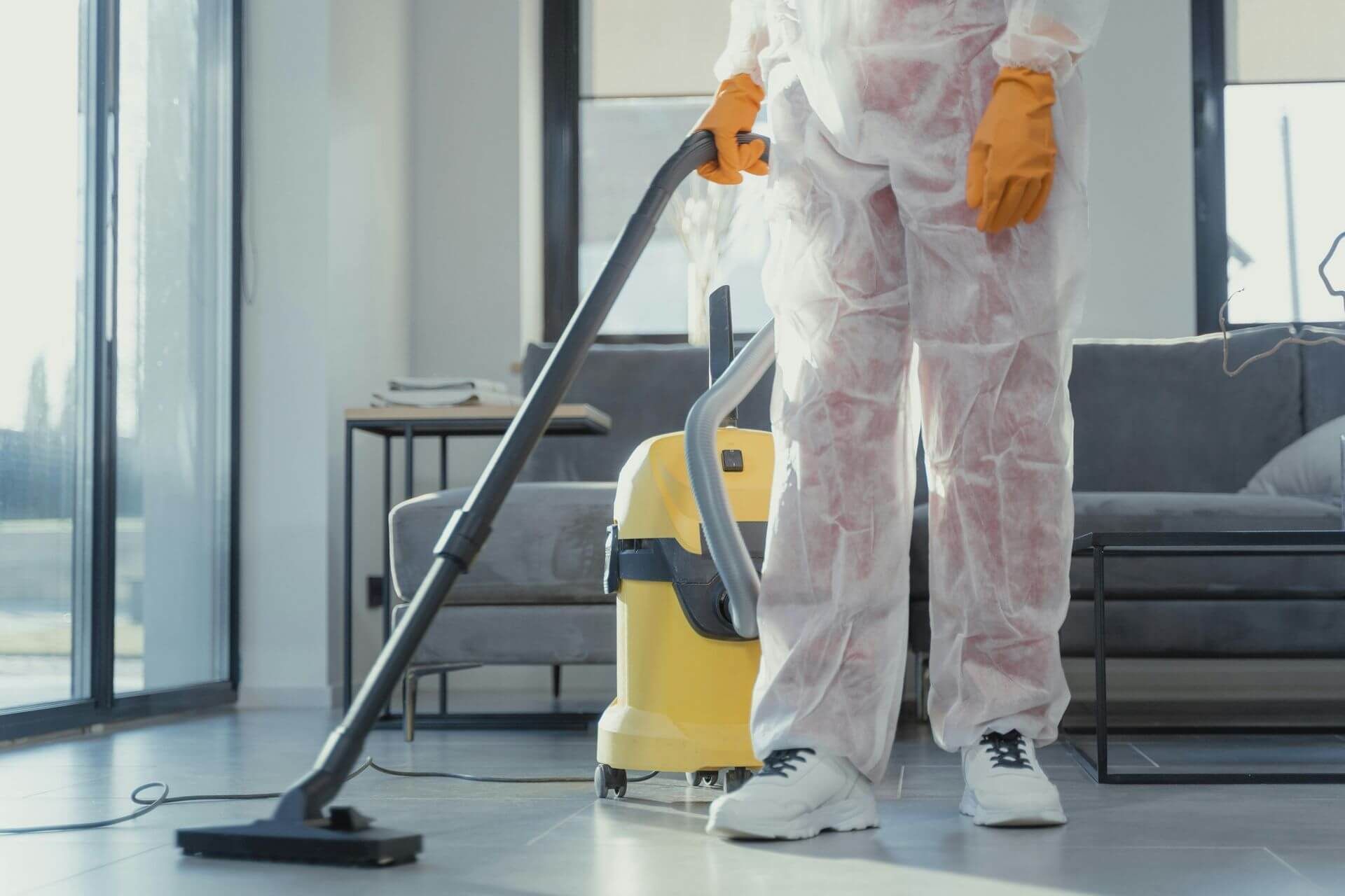 Person in protective suit vacuums a sunlit living room. Yellow vacuum cleaner, orange gloves.