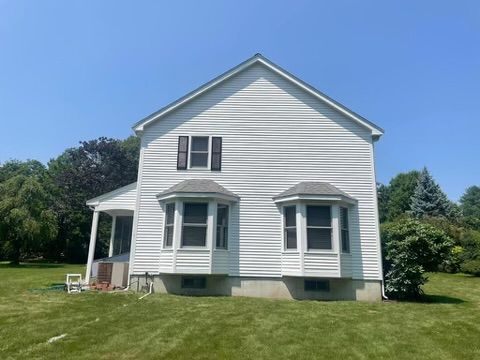 A white house with a large porch is sitting in the middle of a lush green field.