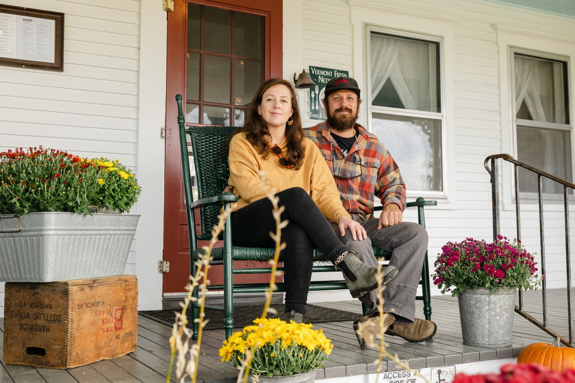 Couple on a porch, sitting on a rocking chair, surrounded by mums and pumpkins.