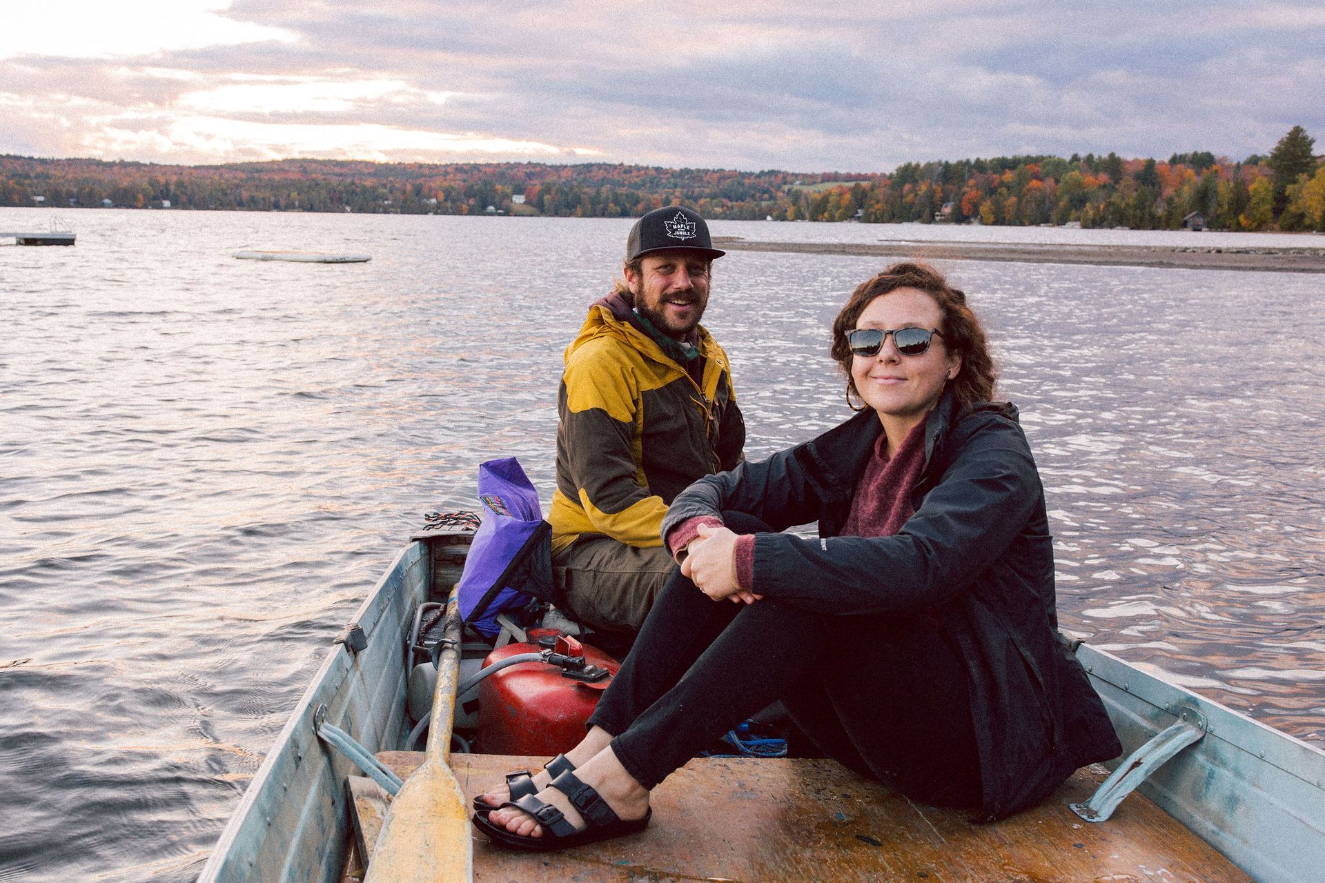 Two people in a metal boat on a lake. The woman wears sunglasses, the man a cap. Autumn foliage in the background.