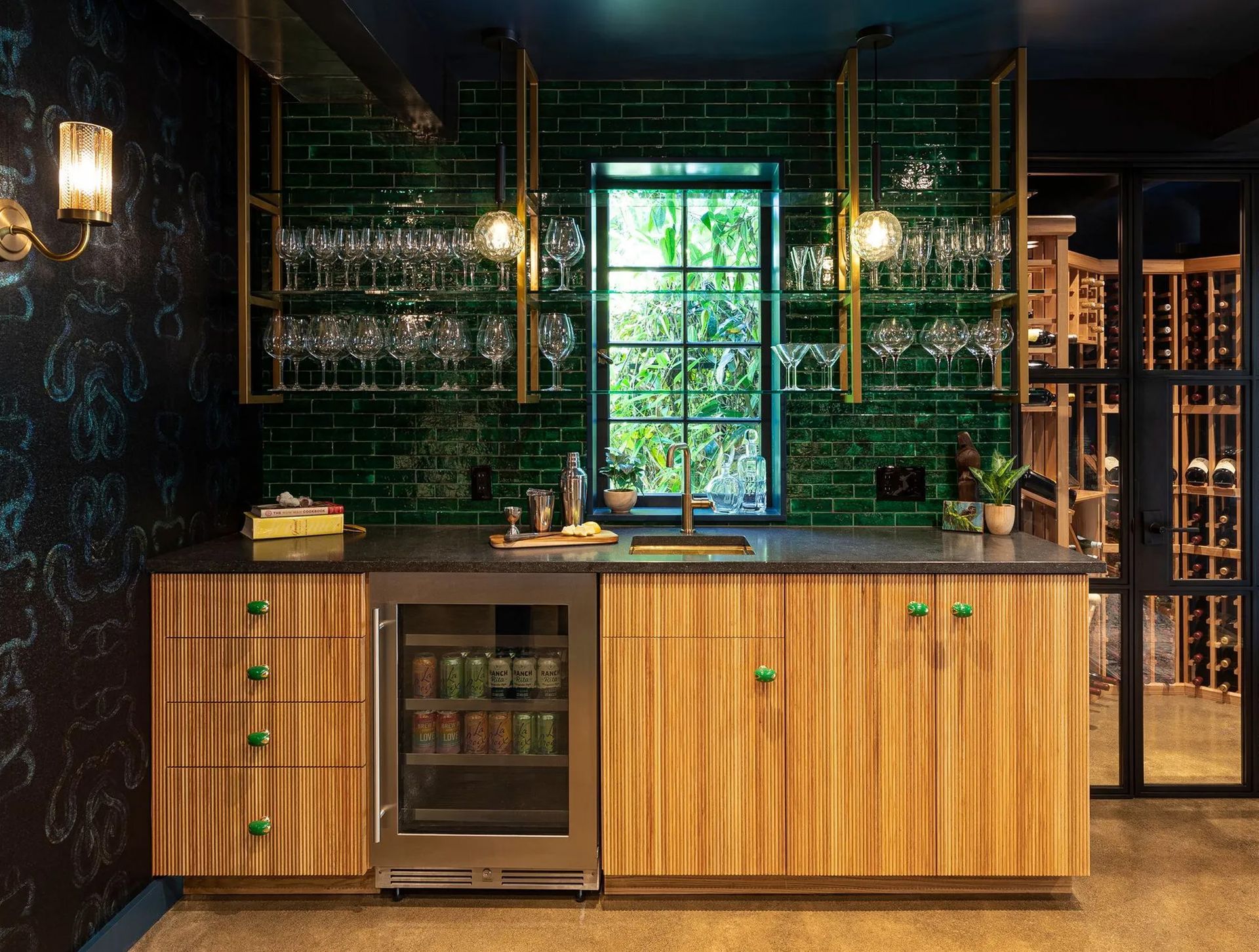 Luxe elements, like brass and glass shelving, reeded wood cabinets, soapstone counters, and glossy green tile make the wet bar the focal point of the newly transformed space. 