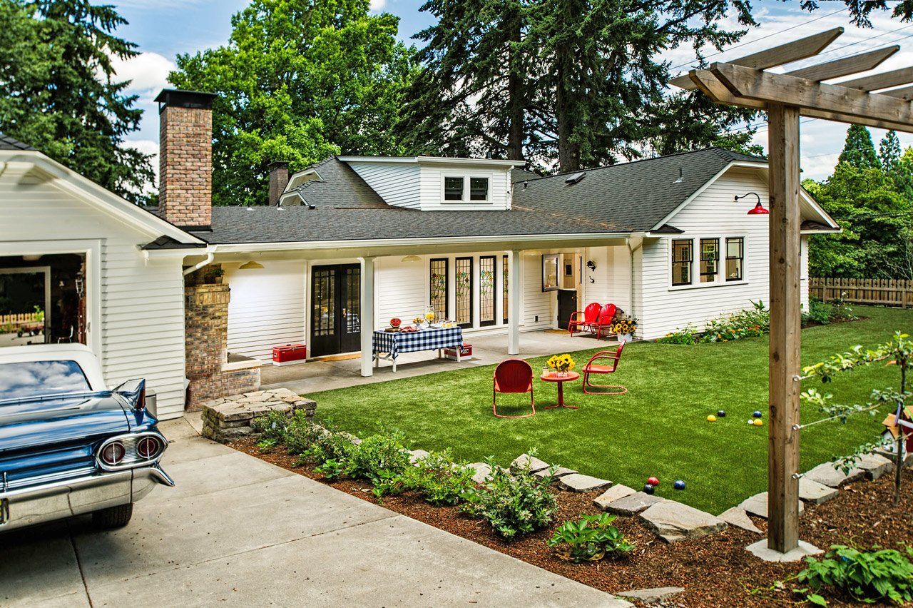 Back of the 1920s home with the garage, breezeway, dormer and back patio with a fireplace visible past the lawn.