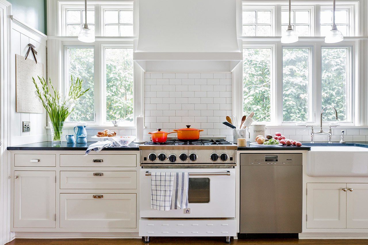Wide view of the kitchen's range wall. New white cabinets and wall tile with a white and stainless steel range sitting alongside a stainless steel dishwasher. Plentiful windows flank the range hood and bring in lots of light.