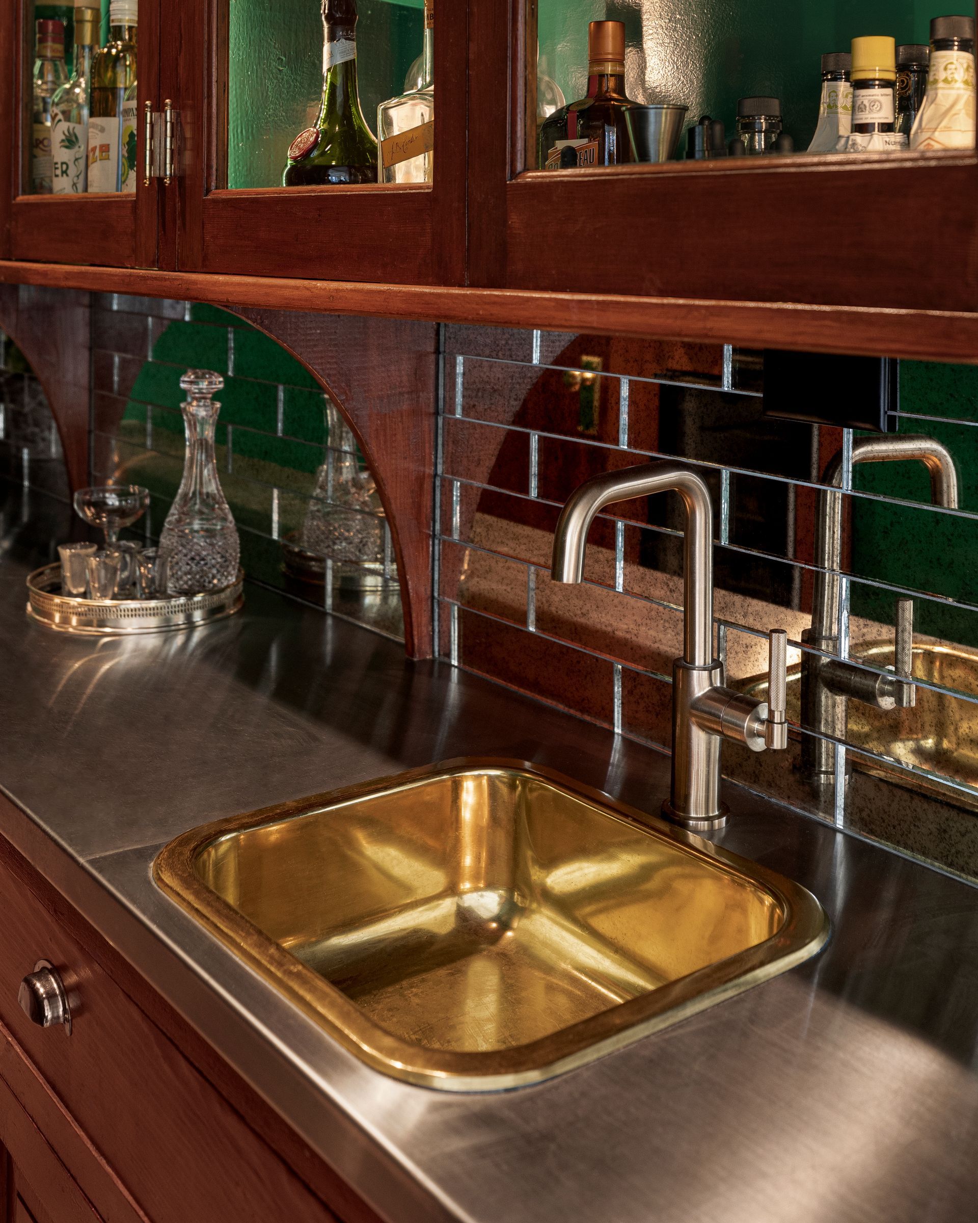 Stainless steel countertops, a brass sink, and a mirrored tile backsplash transform the space into a stylish modern bar. 