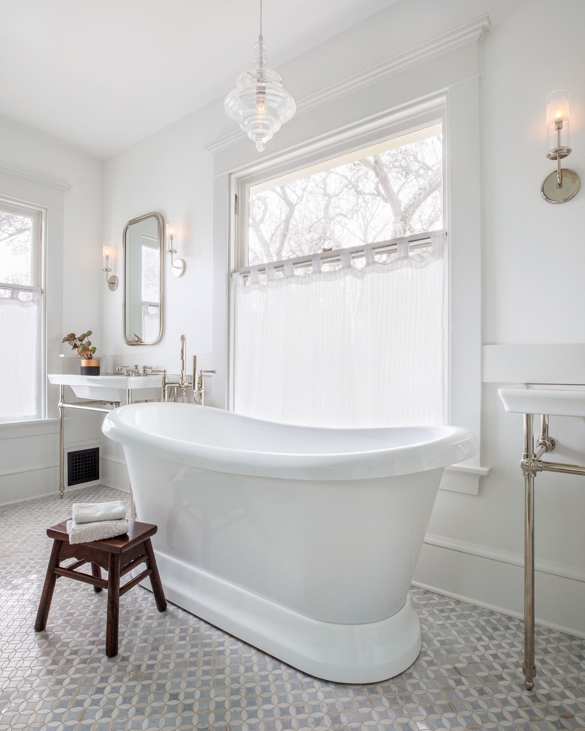 An airy bathroom within a former bedroom features console sinks, a freestanding tub and marble mosaic floors. 