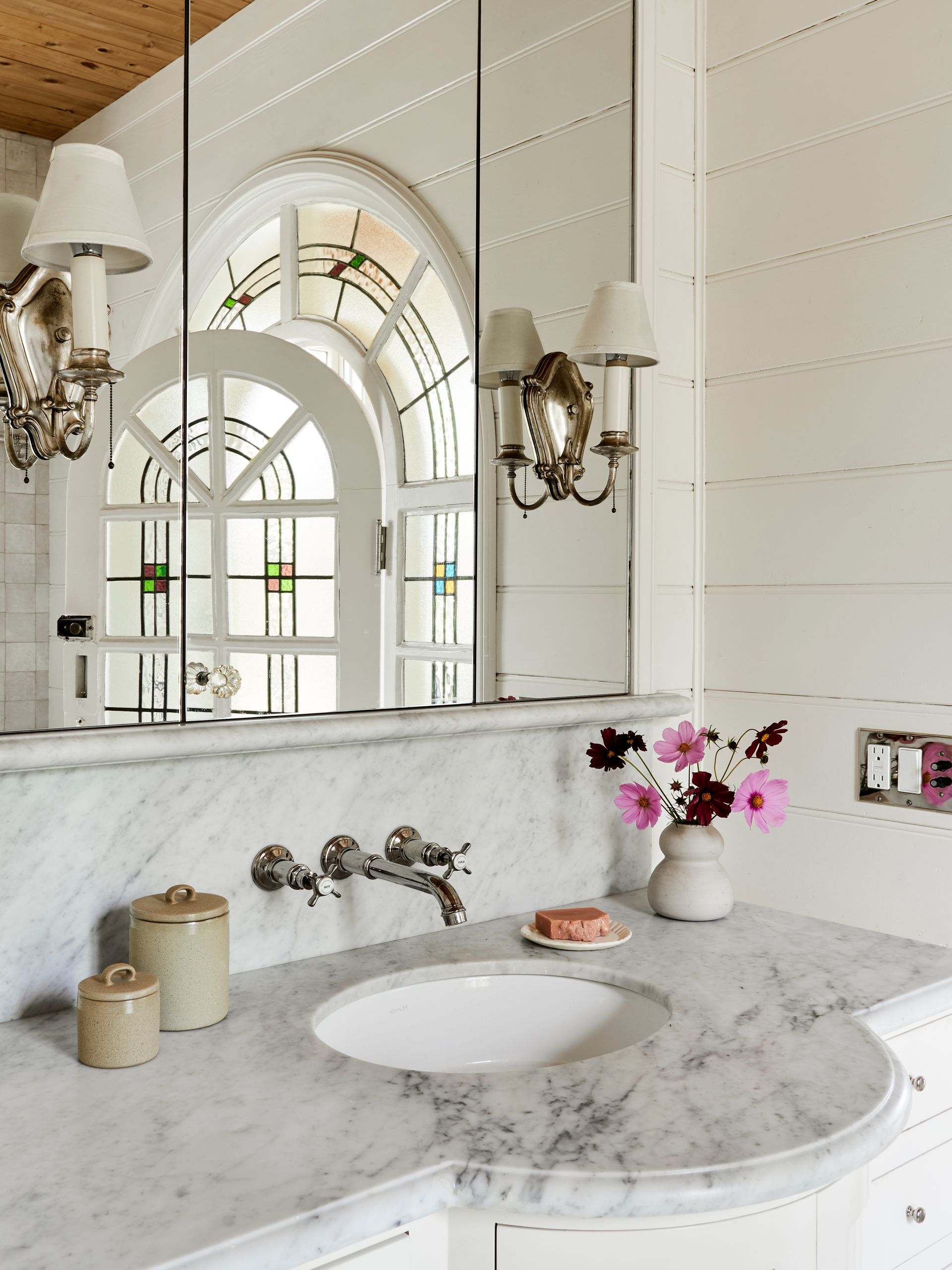This elegant marble-topped vanity is set off by wide beadboard paneling, vintage sconces, and a dramatic salvaged arch-top door.