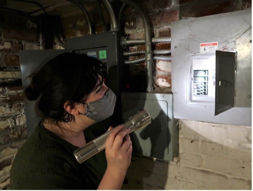 A woman wearing a mask is looking at an electrical box with a flashlight.