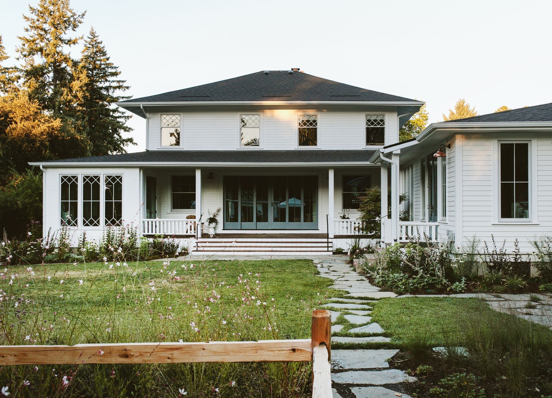 Side of 1910 farmhouse with sunroom and porch addition, large accordion door, cottage style double hung windows with diamond shaped divided lights, a path and flower beds.