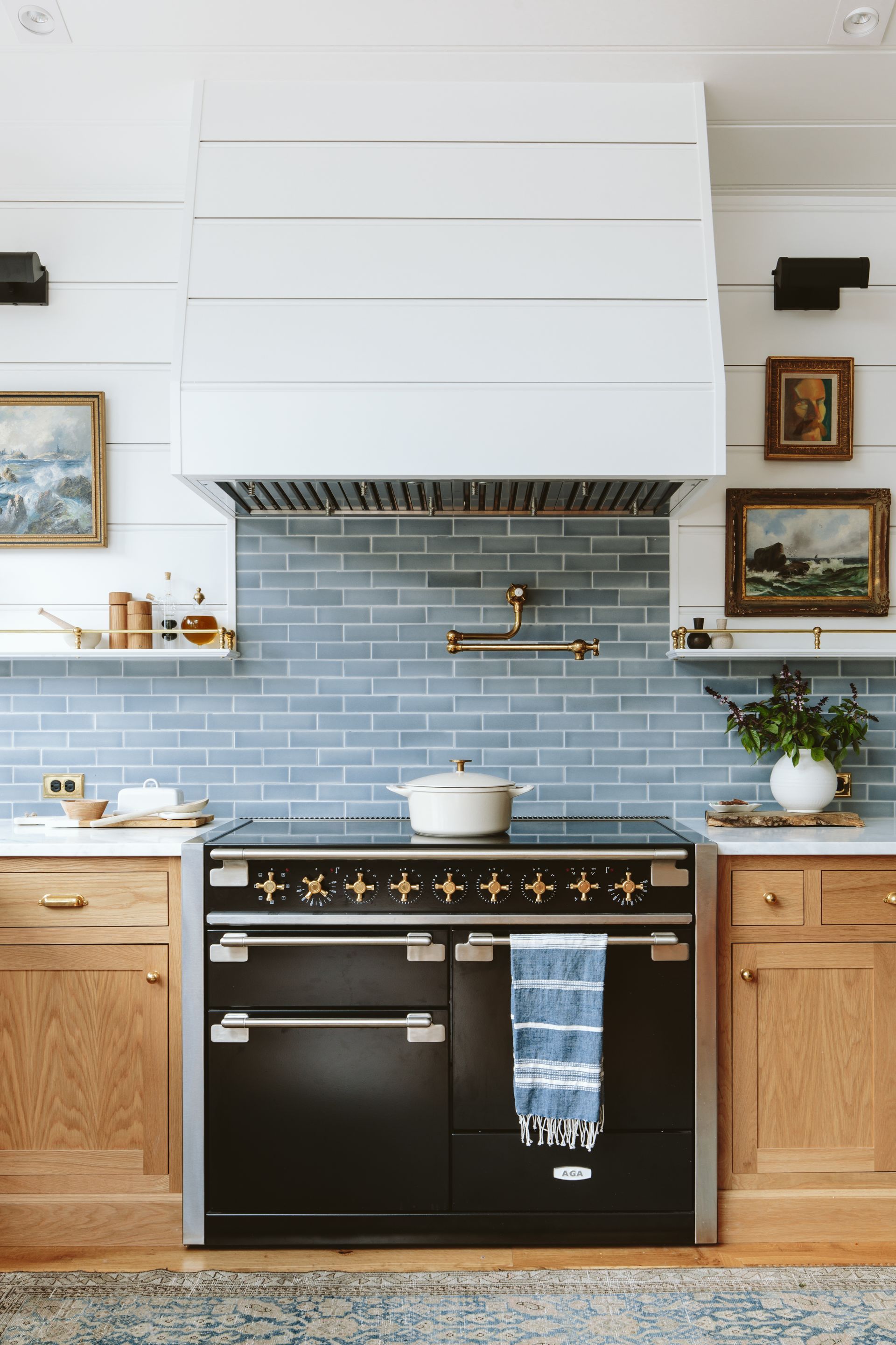 Range wall with custom oak cabinets and large beadboard paneling on the wall and the hood surround, an Aga range, pot filler, Carrara counters, blue tile backsplash capped with a  wooden shelf.