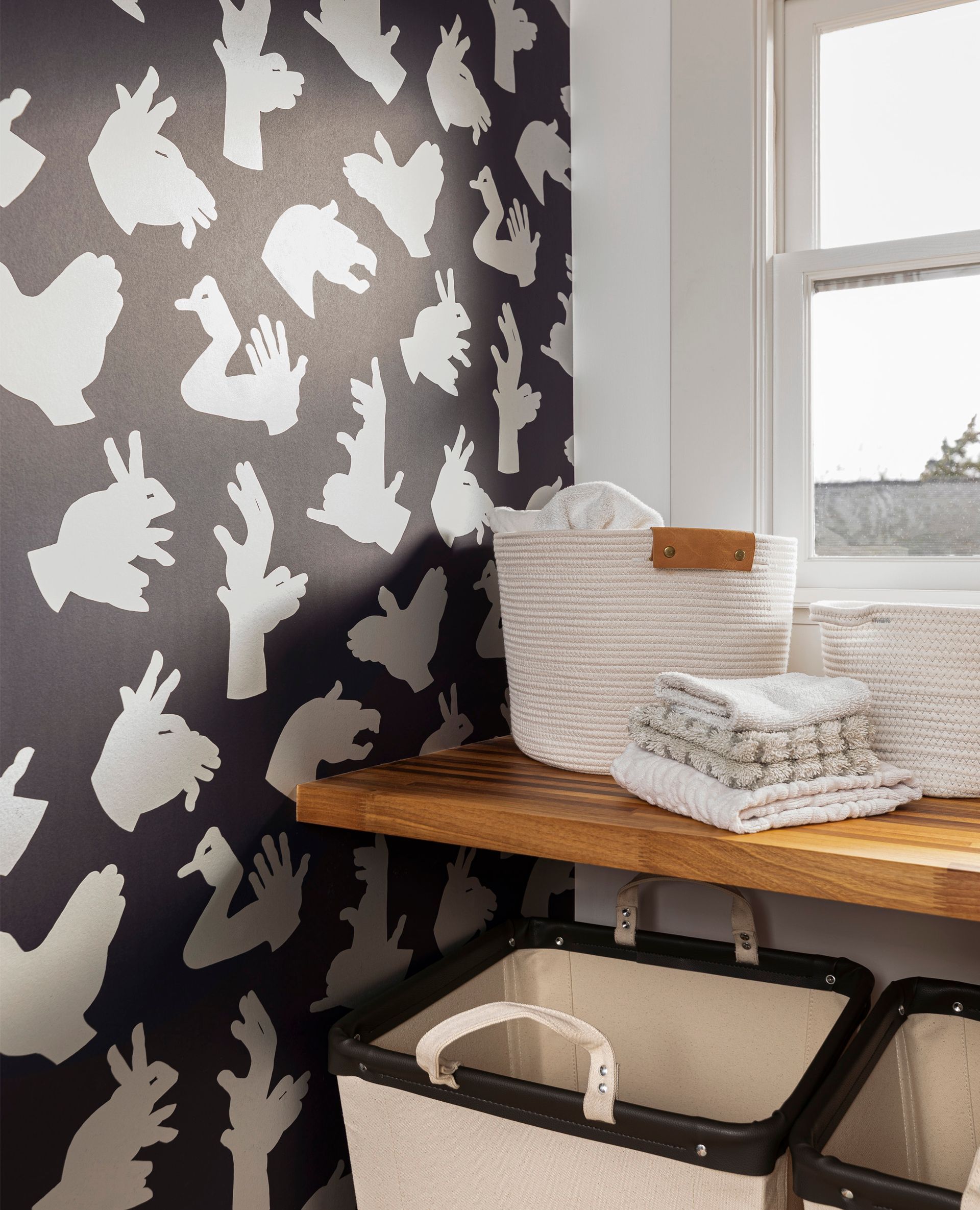 The corner of the laundry room shows shadow puppet wallpaper, a butcher block counter and fabric laundry bins. 