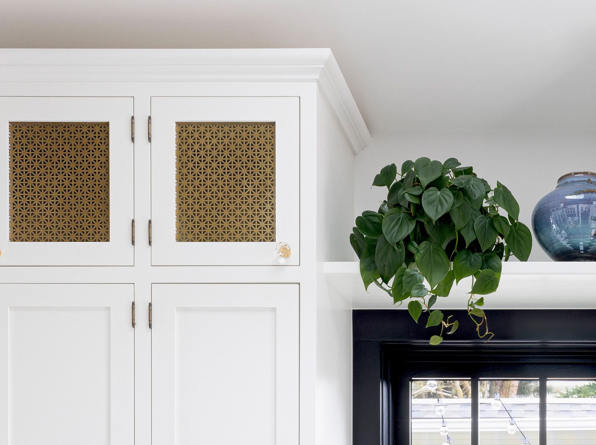 A detail of the upper cabinets, with their brass metal mesh in a decorative pattern. Adjacent, there's a shelf for plants and treasured ceramics above the window. 