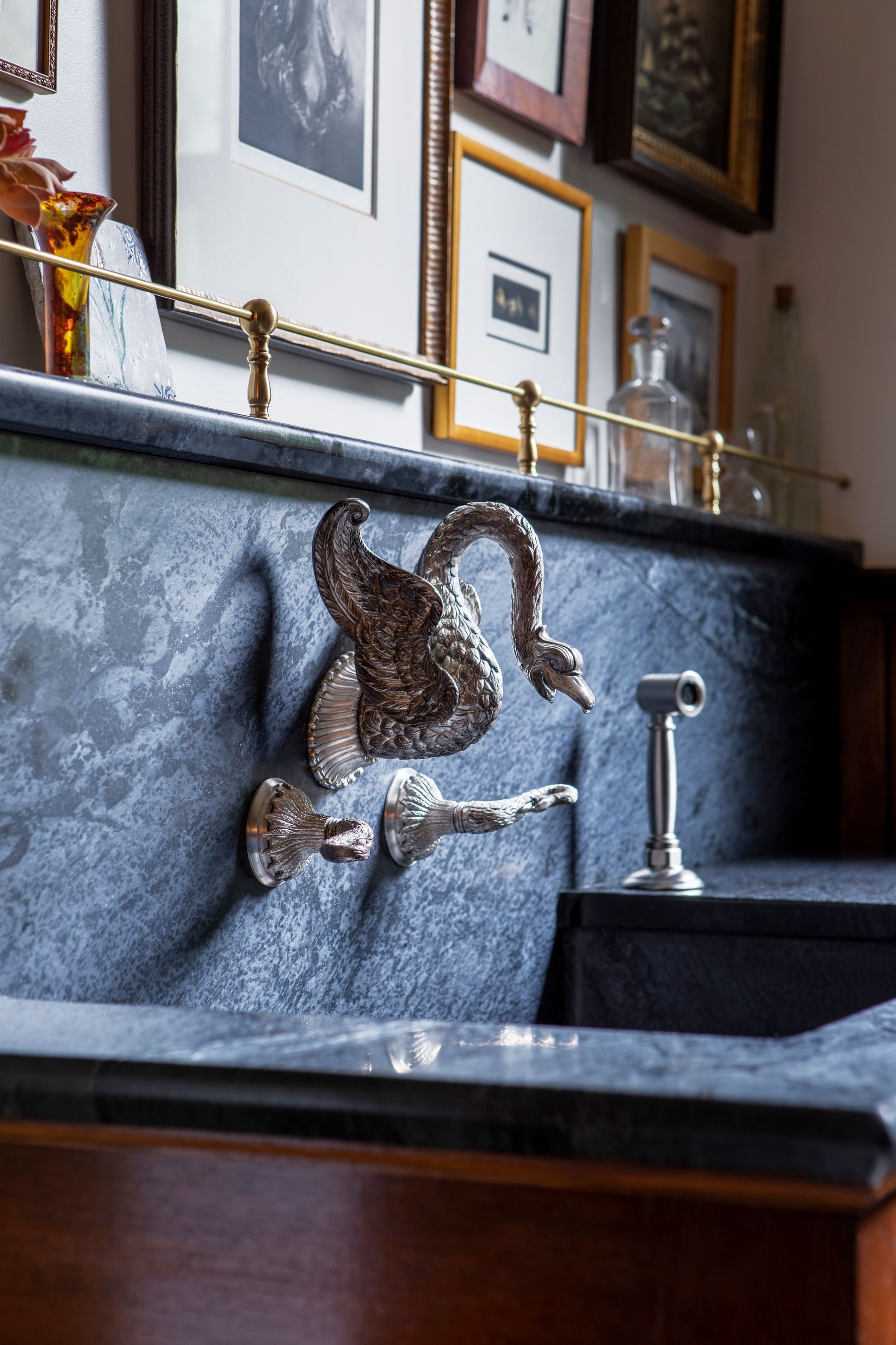 Close up of a custom soapstone sink, vintage wall mounted swan faucet, and integrated backsplash below a picture shelf with brass rails and eclectic artwork.