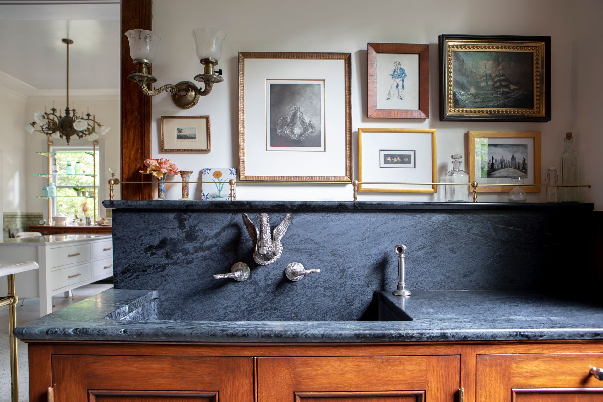 Custom soapstone sink with vintage wall mounted swan faucet with integrated backsplash below a picture shelf with brass rails and eclectic artwork.