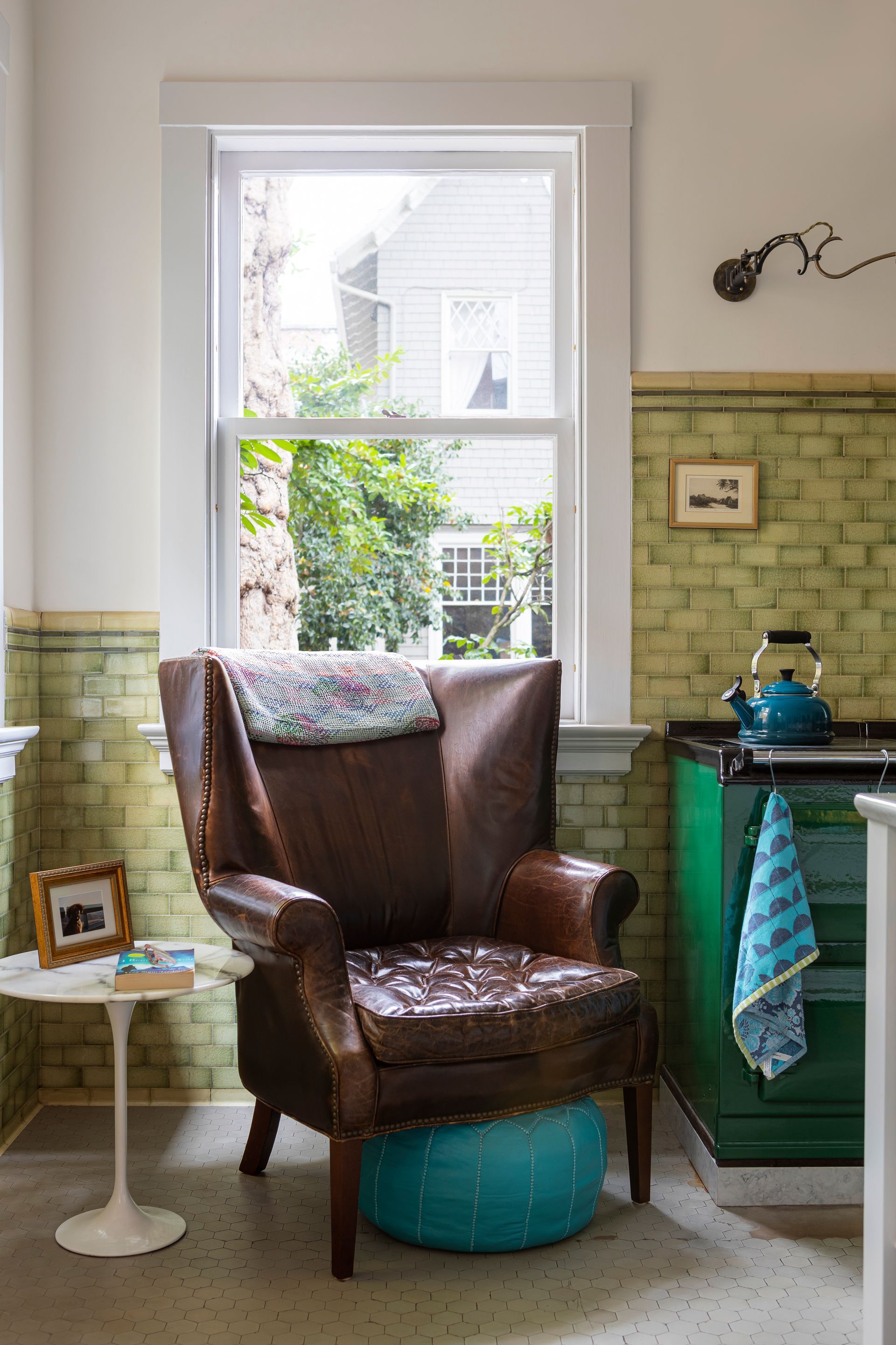 Cozy kitchen corner with leather wingback chair next to warm vintage Aga range in front of original celadon colored subway tile.