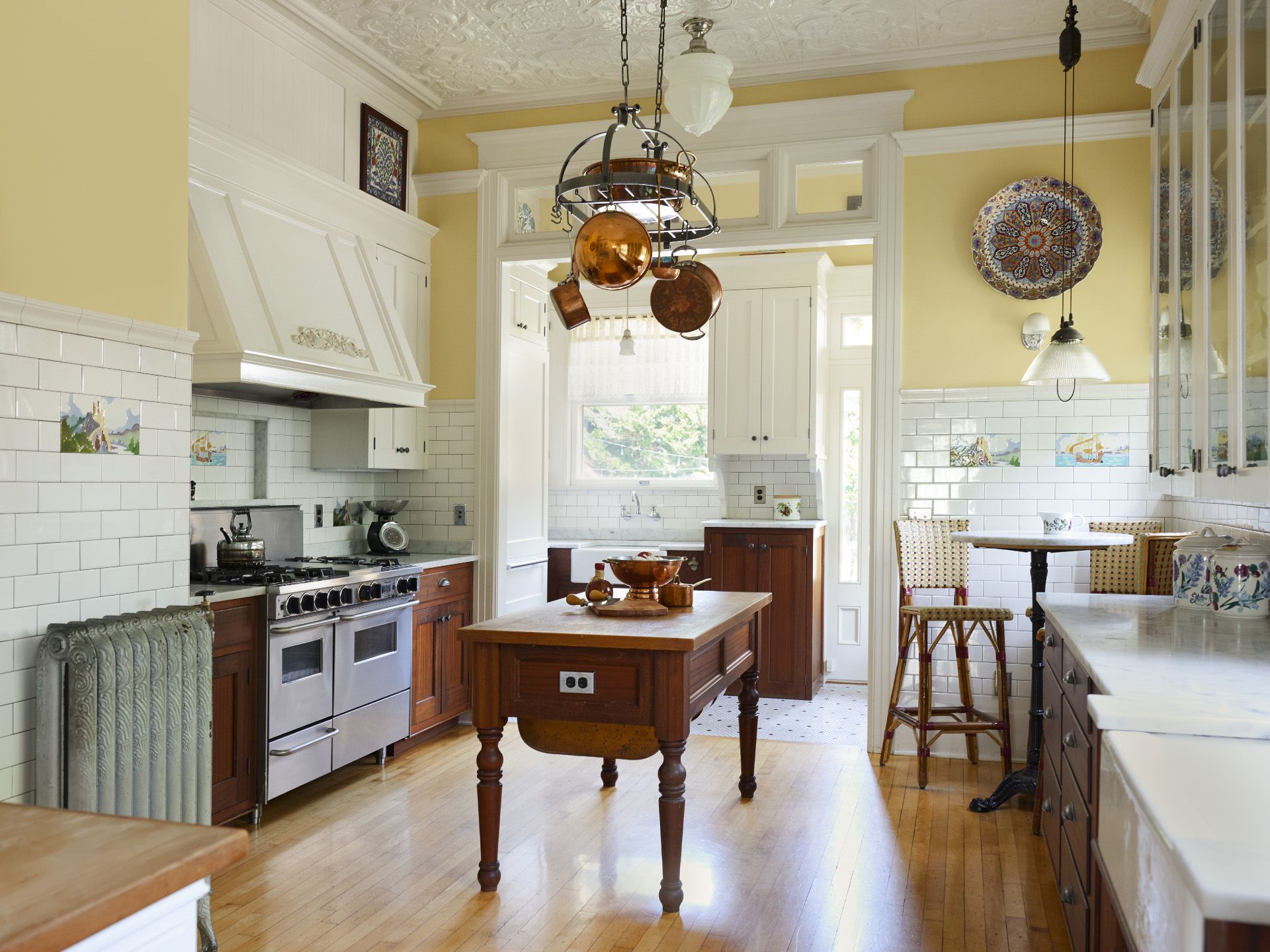 The kitchen evokes a French bakery feel with high ceilings clad in painted pressed tin, butter yellow walls and fresh white tile as wainscoting. Base cabinets and a central island are stained mahogany and the range hood and trim are painted white.