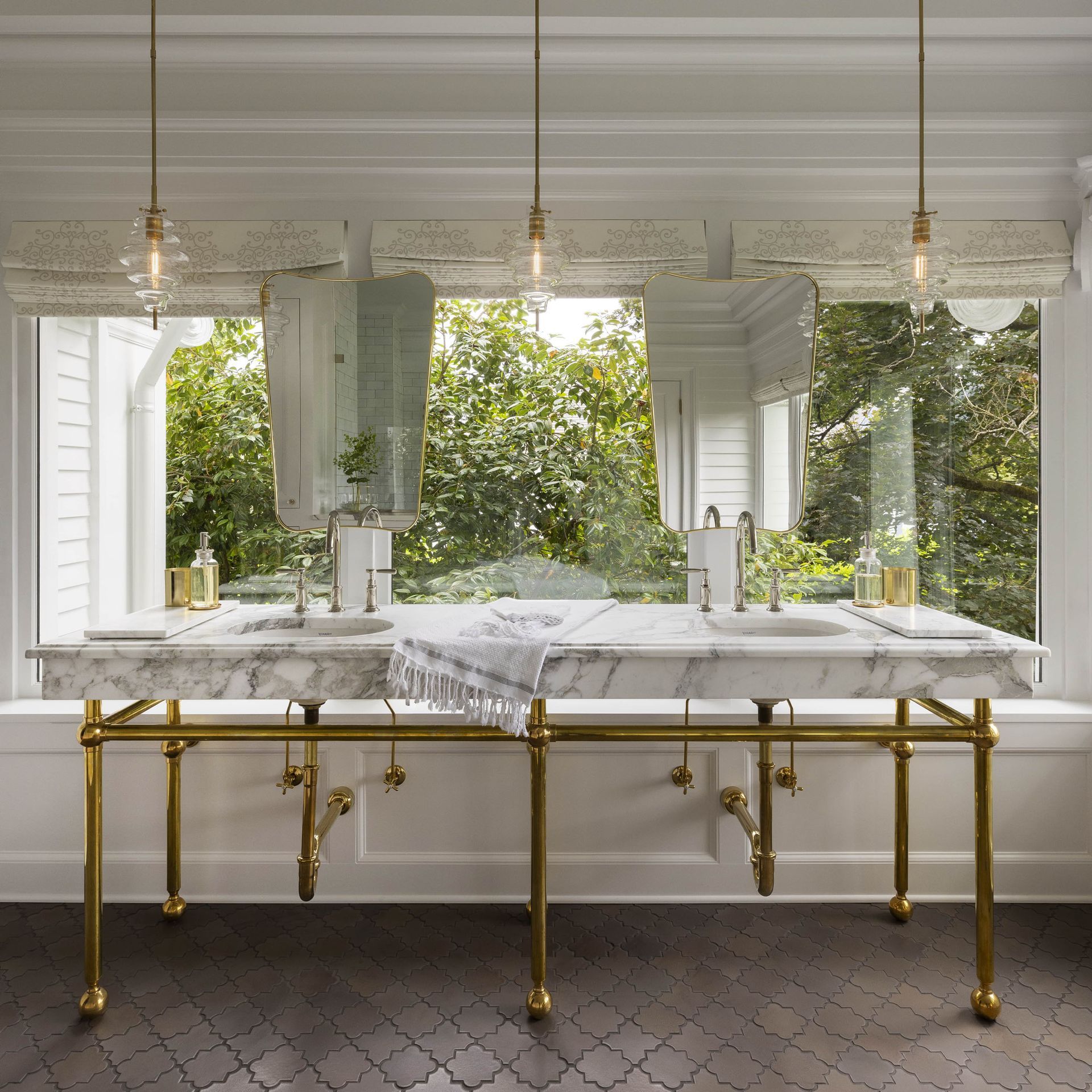 A custom double vanity with marble counters and apron, a brass console base, mirrors mounted in front of window mullions and pendants with clear glass shades in this main bath.