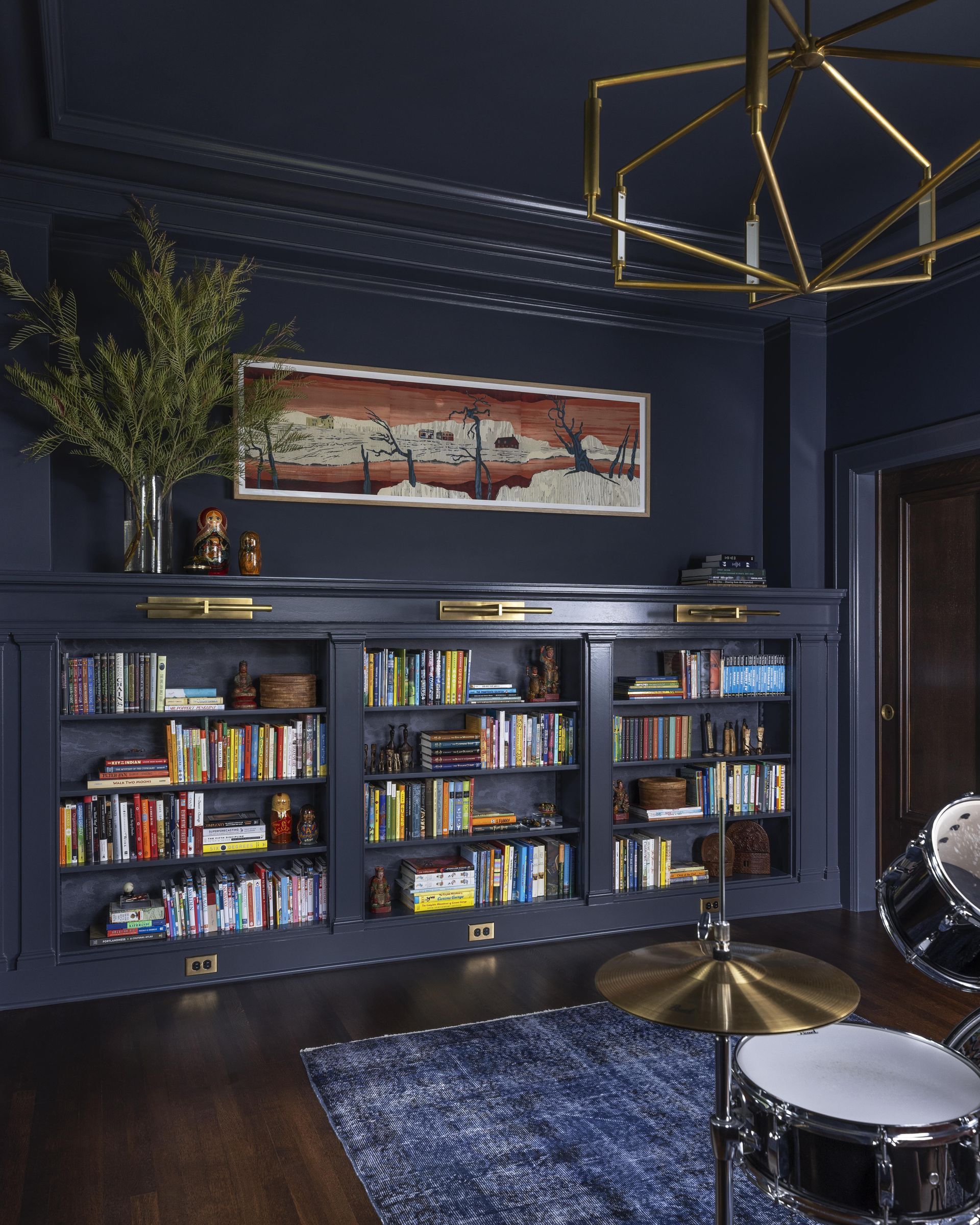 A deep, dark blue den features a drum set, Kelly Wearstler light fixtures and dark stained oak floors.