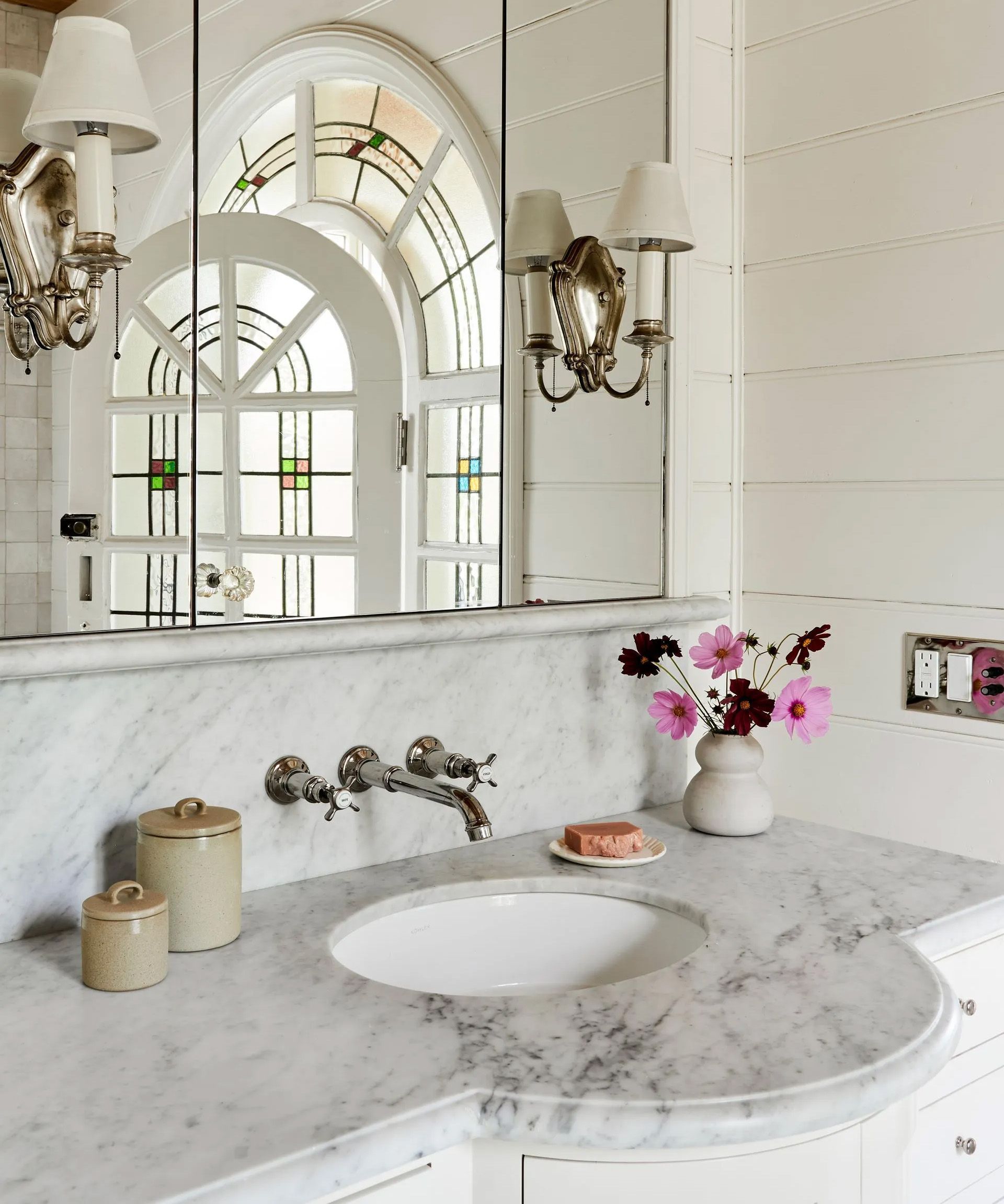 This elegant marble-topped vanity is set off by wide beadboard paneling, vintage sconces, and a dramatic salvaged arch-top door. 