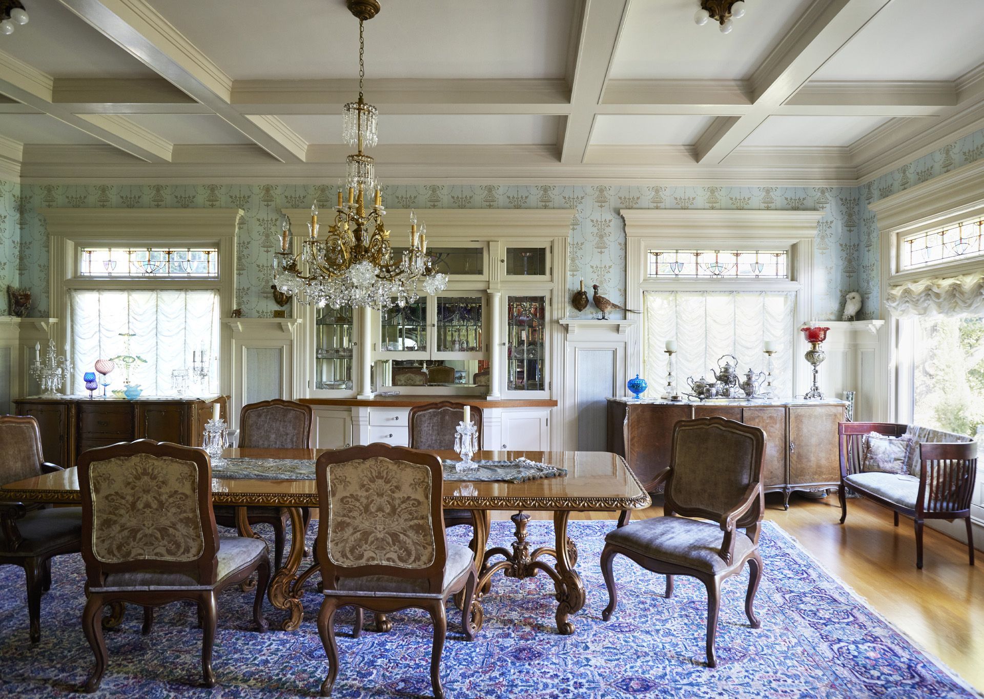 The formal dining room has painted millwork and robins egg blue wallpaper with gold accents. The built in, flanked by large windows, has leaded glass doors above the counter to show off favorite servewear.